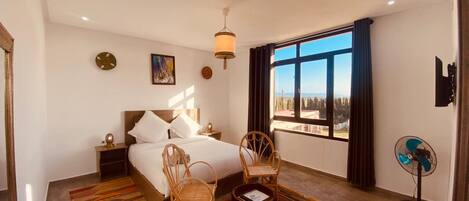 A bedroom with a large bed, two chairs, a small table, a standing fan, and a window with a view.