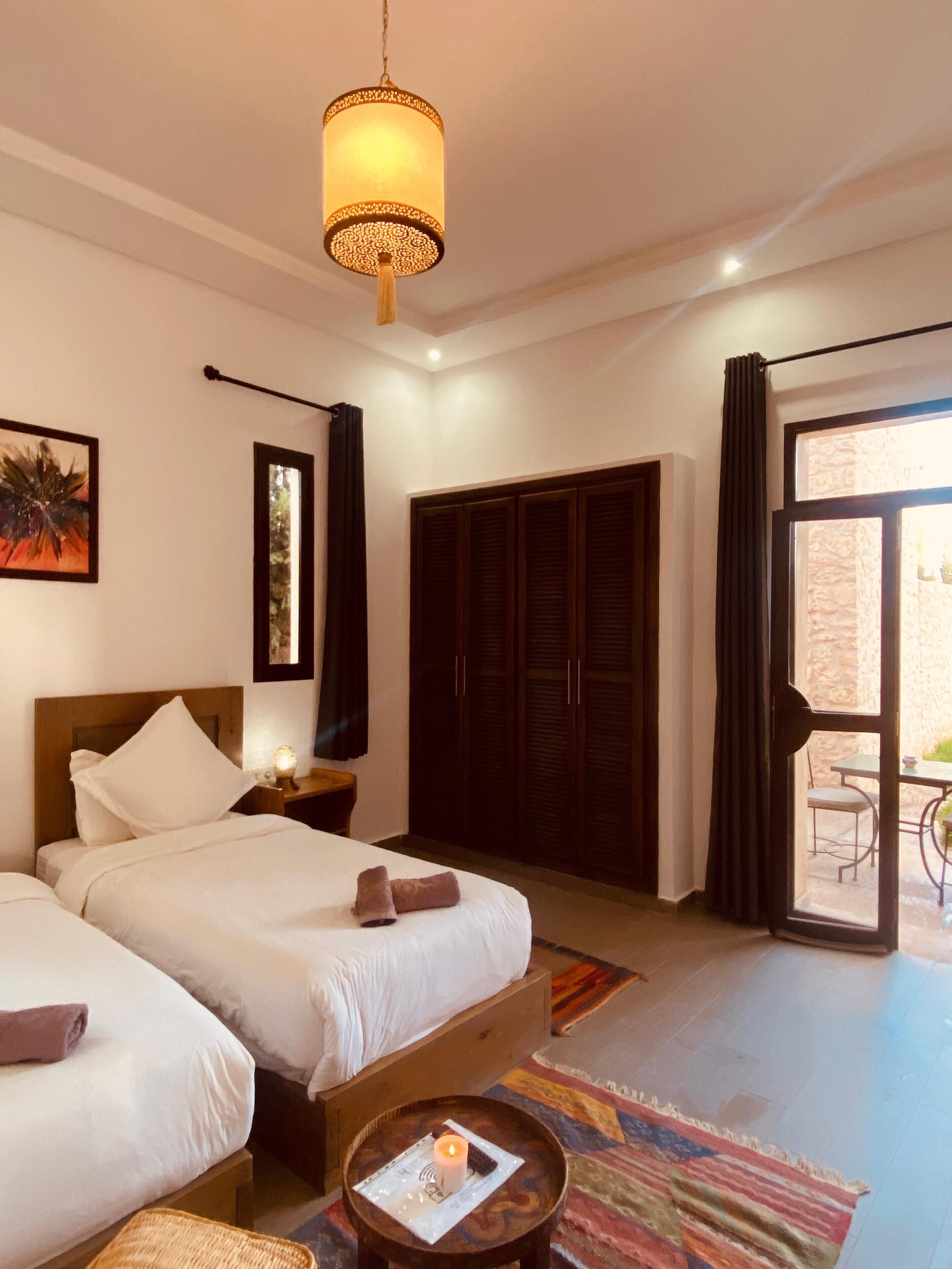 A hotel room with two beds, a large wardrobe, a dining area with a table and chairs, and a sliding glass door to the outside.