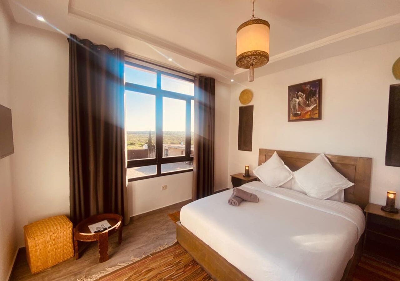 A hotel room with a large bed, a view through the window, a small table, and a hanging light fixture.