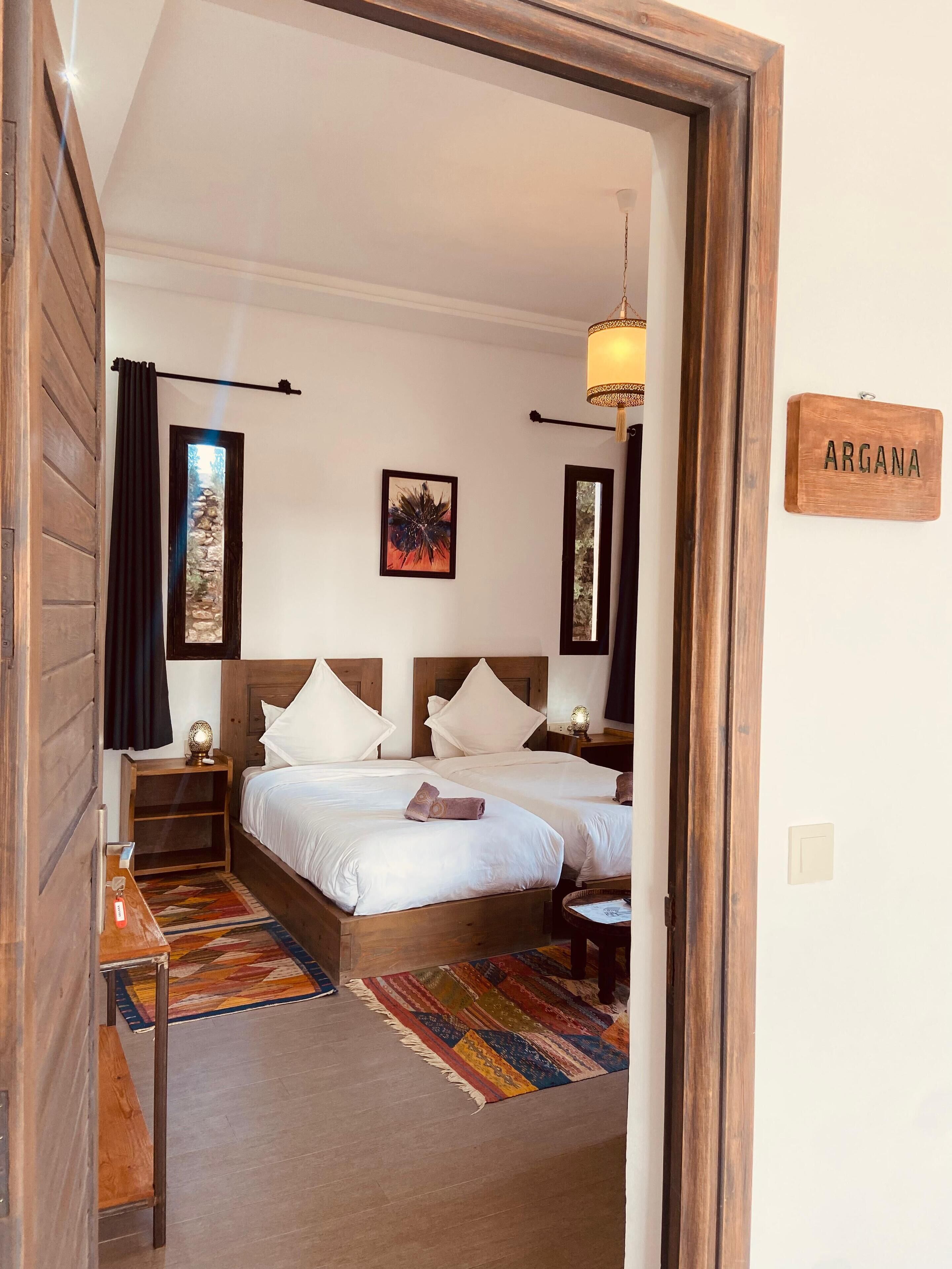 Un dormitorio con dos camas, cabecero de madera, mesita de noche, lámpara, alfombra y un cartel que dice "ARGANA".