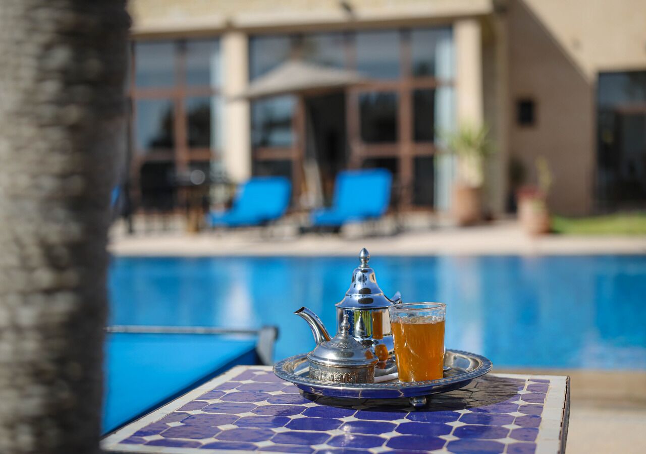 A poolside table with a tray containing a teapot and a glass of tea, set against a backdrop of a swimming pool and a building with large windows and blue chairs.