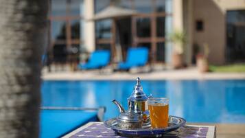 A poolside table with a tray containing a teapot and a glass of tea, set against a backdrop of a swimming pool and a building with large windows and blue chairs.