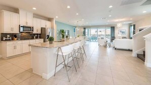 Una cucina moderna con mobili bianchi, un piano di lavoro in granito ed elettrodomestici in acciaio inossidabile. È presente una zona bar con sgabelli e un'area pranzo con un divano bianco e un lampadario.