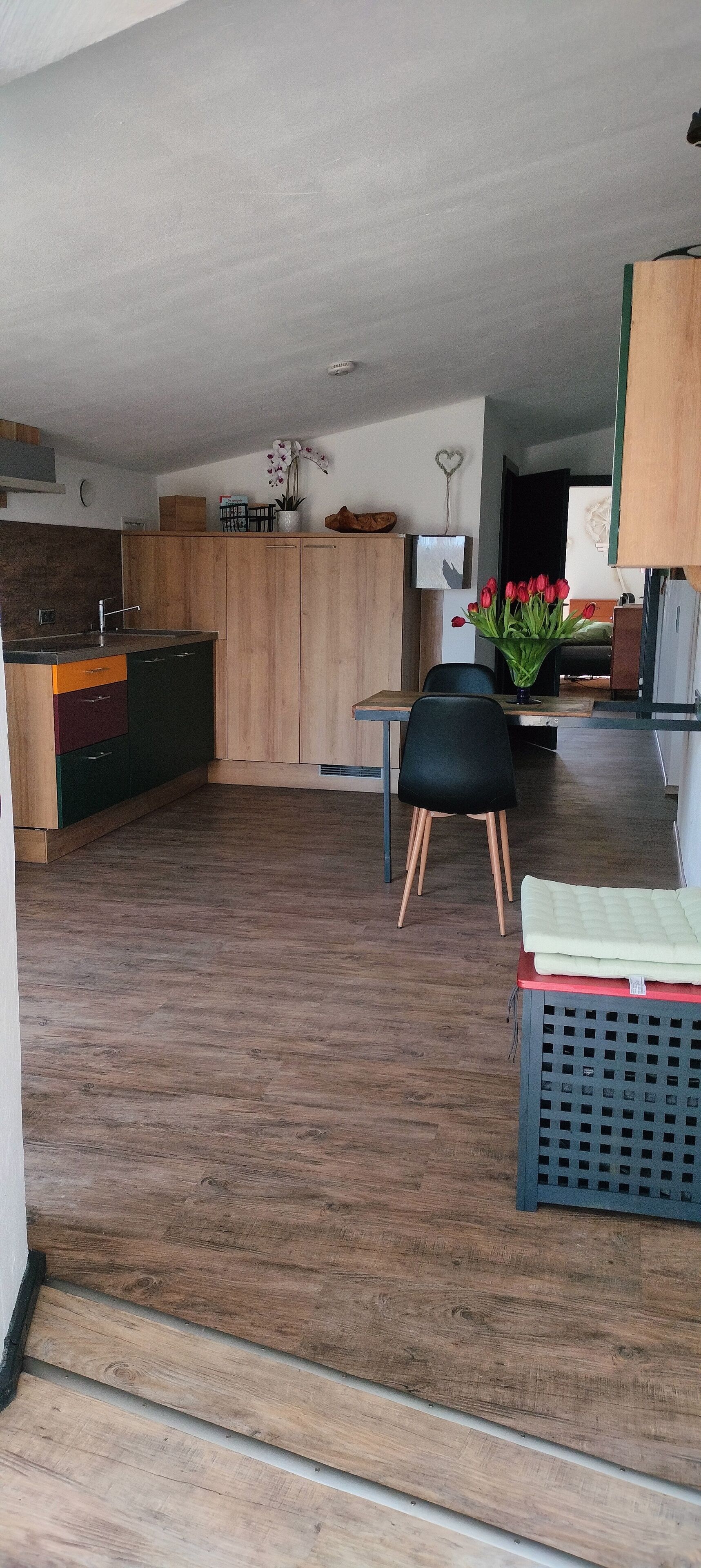 Moderni keittiö, jossa on puiset kaapit, ruokailutila, jossa on musta tuoli, ja pöydällä oleva maljakko täynnä punaisia kukkia.