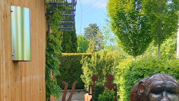 Een smal pad begrensd door houten muren en een metalen poort, leidend naar een tuin met een groot metalen sculptuur en diverse planten.