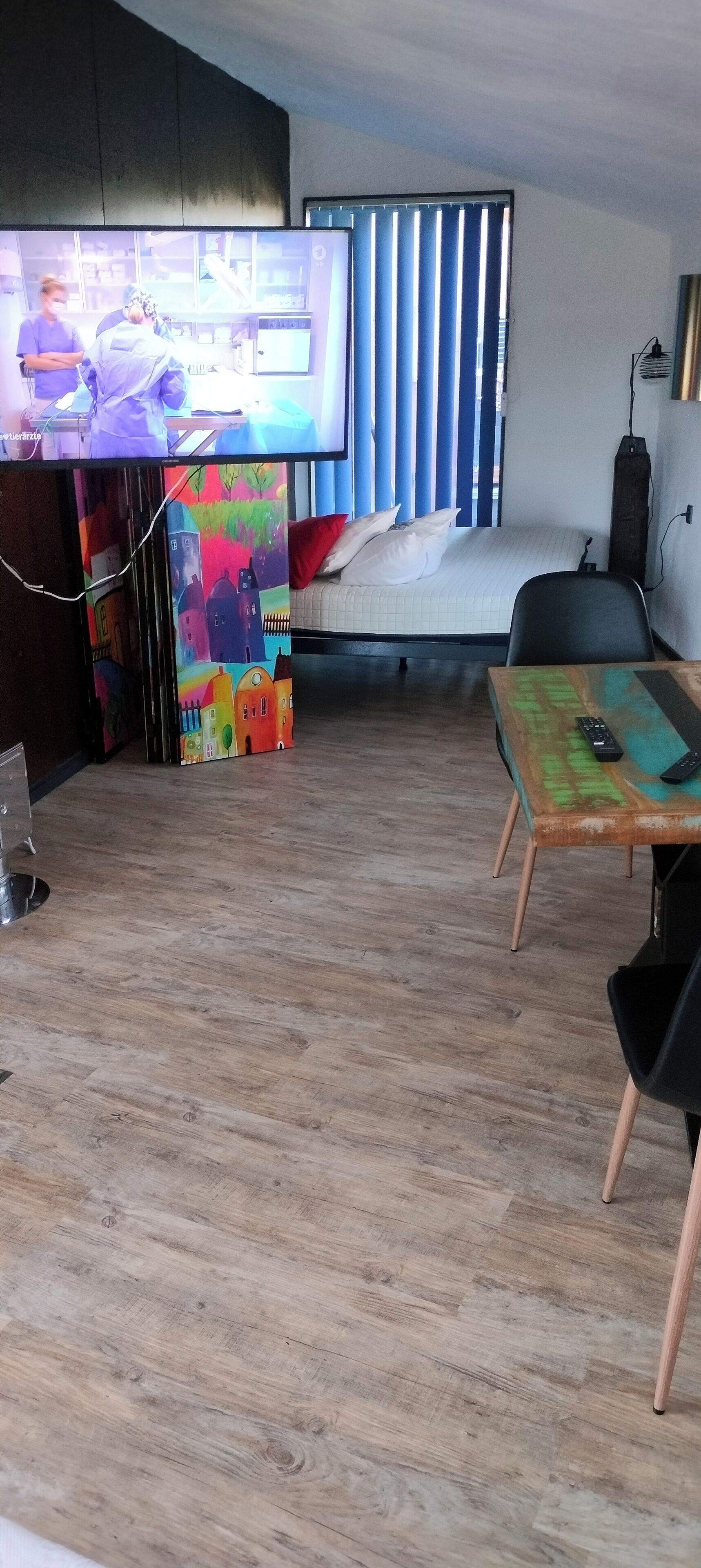 Une pièce avec un plancher en bois, un lit recouvert de draps blancs, un téléviseur fixé au mur et une fresque murale aux couleurs vives.