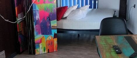 Une pièce avec un plancher en bois, un lit recouvert de draps blancs, un téléviseur fixé au mur et une fresque murale aux couleurs vives.