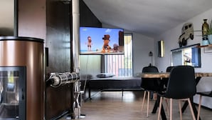 Een moderne woonkamer met een grote televisie, een koffieapparaat, een eethoek met stoelen en een plant aan de muur.