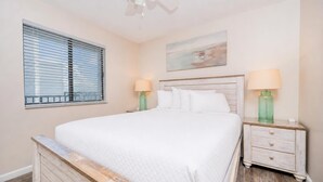 A bedroom with a bed, two bedside tables, a window with blinds, a ceiling fan, and a painting on the wall. - Gulf Shores Surf & Racquet 614a 1 Bedroom Condo (Gulf Shores)