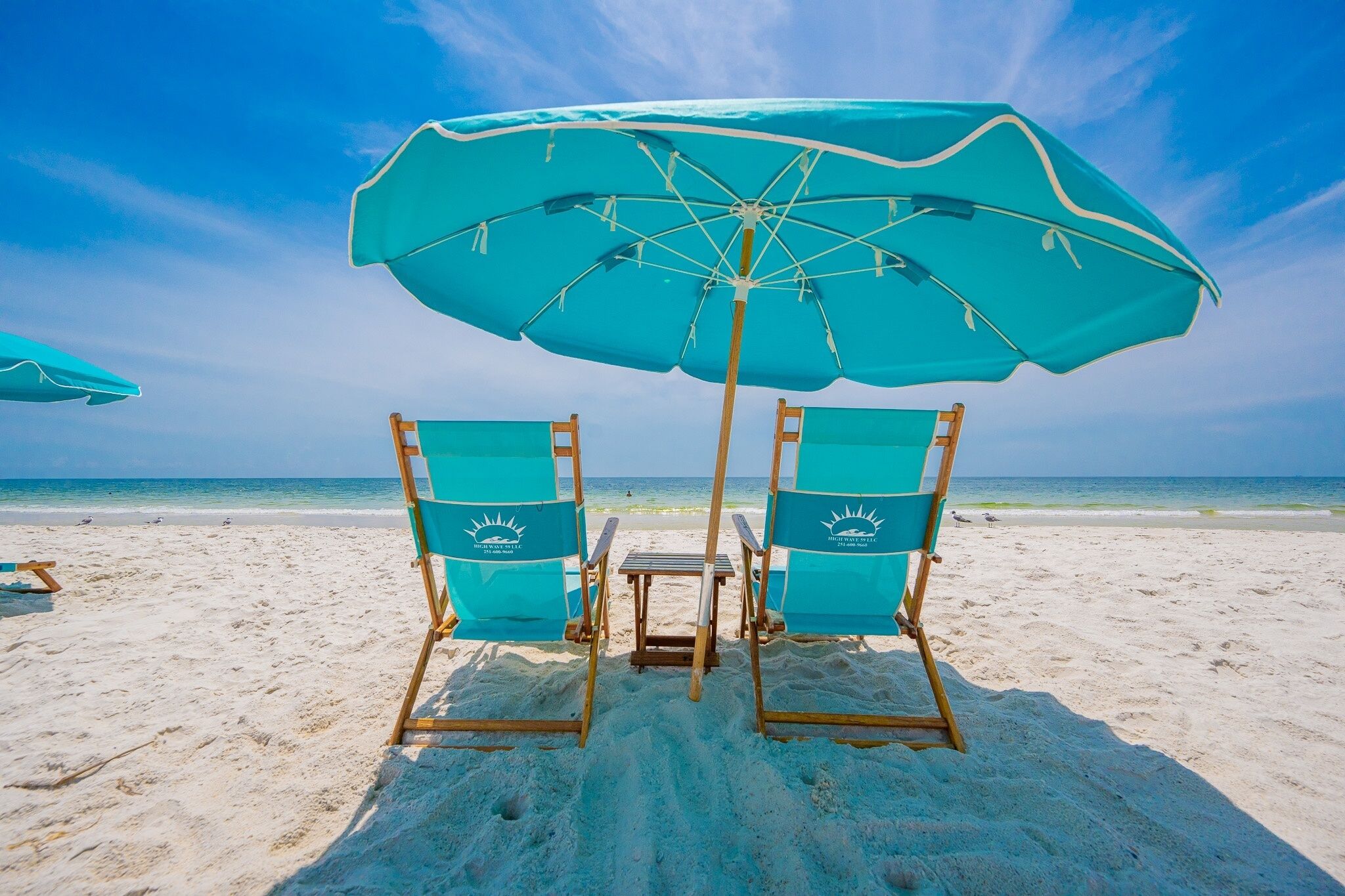 Two beach chairs with a table under a large umbrella on a sandy beach.