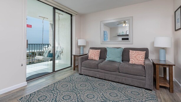 A living room with a grey sofa, patterned rug, and two side tables with lamps. A large window offers a view of a balcony with outdoor furniture. - Gulf Shores Surf & Racquet 614a 1 Bedroom Condo (Gulf Shores)