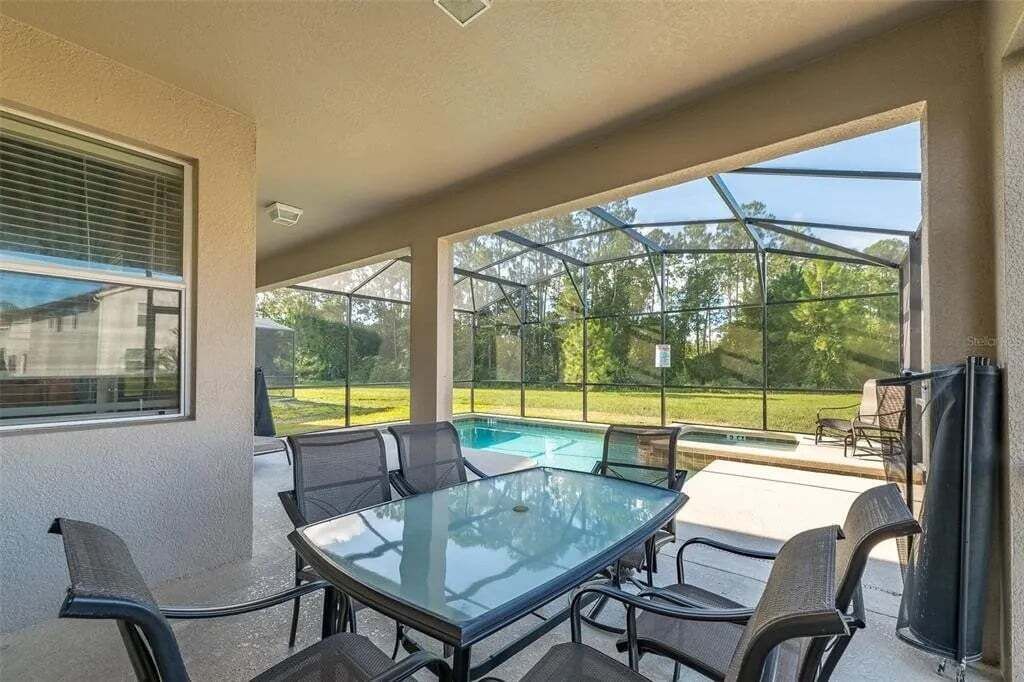 A covered patio with a glass table and chairs, overlooking a swimming pool and a grassy yard.