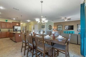 A spacious dining area with a wooden table set for six, surrounded by chairs. A chandelier hangs above, and a ceiling fan is visible. The kitchen area is visible in the background with wooden cabinets and a refrigerator.