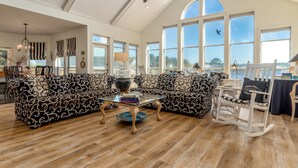 A spacious living room with a large sectional sofa, a glass coffee table, and a wooden rocking chair. - At Ease - 535 Barcelona 3 Bedroom Home by RedAwning (Dauphin Island)