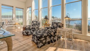 A sunlit room with large windows, a white bench, patterned ottomans, a small table, and a lamp. - At Ease - 535 Barcelona 3 Bedroom Home by RedAwning (Dauphin Island)