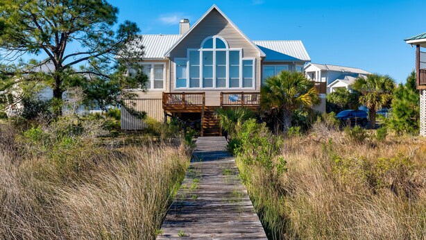 A house with a wooden deck leading to a large front door, surrounded by tall grass and trees. - At Ease - 535 Barcelona 3 Bedroom Home by RedAwning (Dauphin Island)