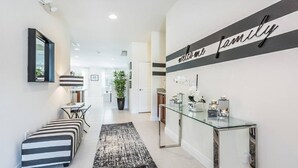 Un couloir moderne avec un banc à rayures, une console en verre agrémentée d’objets décoratifs et un panneau indiquant : « Bienvenue à la famille ».
