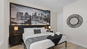 Un dormitorio moderno con una pintura de un paisaje urbano sobre la cama, un espejo redondo en la pared y una cama bien hecha con una manta negra.