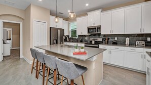 Una cocina moderna con armarios blancos, una isla central con taburetes y una variedad de electrodomésticos, incluyendo un refrigerador, microondas y estufa.