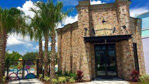 A fitness center with a brick facade, large glass entrance, and palm trees in the foreground. - Last Minute Disney Getaway! Private Pool & Themed Bedrooms. Book Now! #77303 (Davenport)