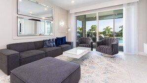A modern living room with a grey sectional sofa, a glass coffee table, and a grey armchair. There is a large window with a view of palm trees and a pool. - Last Minute Disney! Private Pool, Hot Tub, Game Room & Themed Bedrooms. Book Now! #79591 (Orlando)