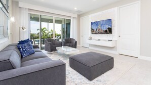 A modern living room with a grey sofa, ottoman, and armchairs, a glass coffee table, and a large flat-screen TV mounted on the wall. - Last Minute Disney! Private Pool, Hot Tub, Game Room & Themed Bedrooms. Book Now! #79591 (Orlando)