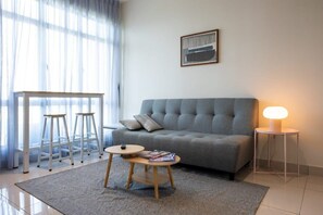 Una sala de estar con un sofá gris, una mesita redonda con una lámpara y un ventanal con cortinas translúcidas.