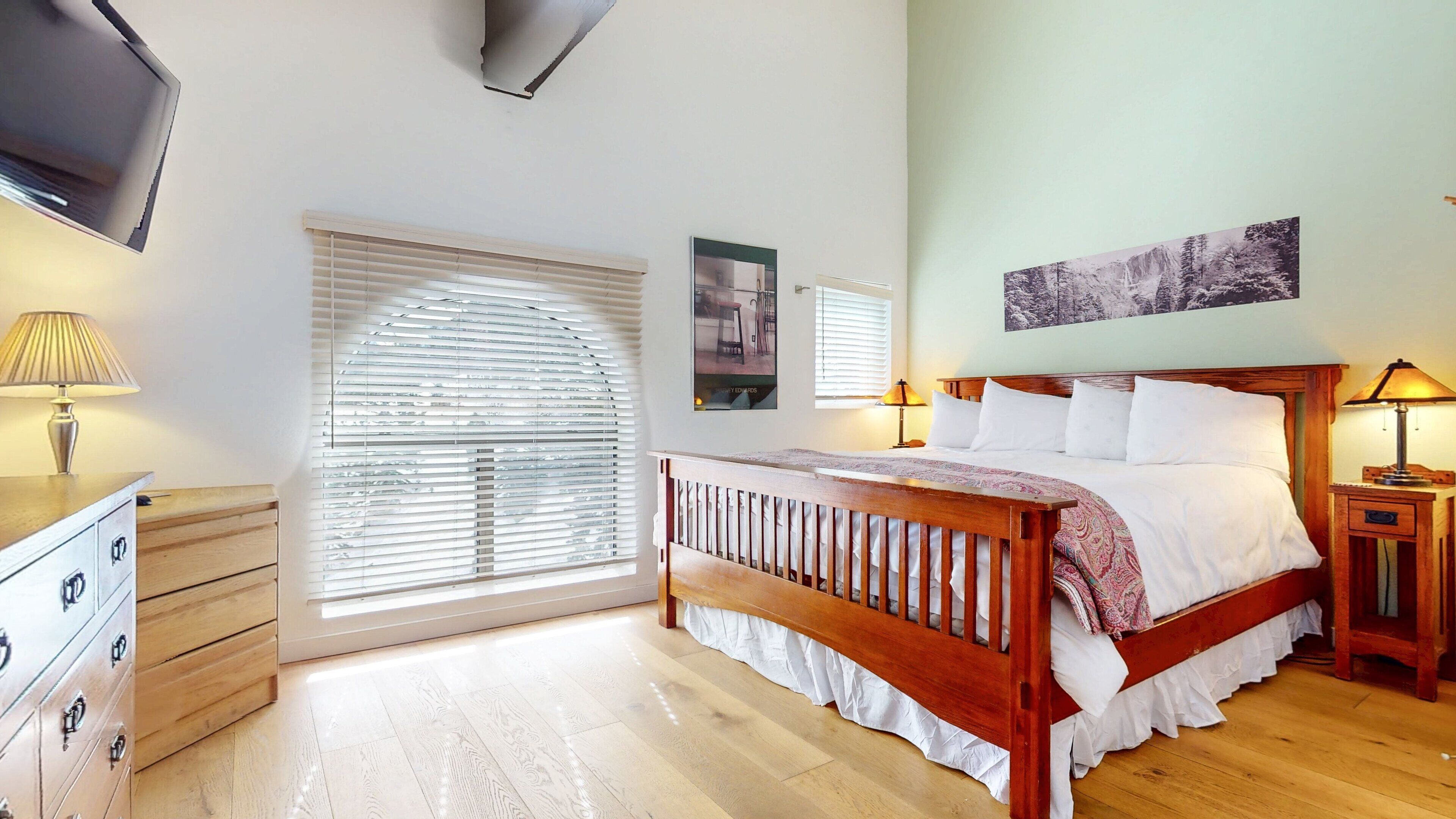 A bedroom with a wooden bed, a dresser, a nightstand, and a TV mounted on the wall.