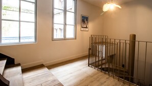 A room with wooden flooring, a staircase with a metal railing, and a window with a view of buildings outside. - Le Coursarlon (Bourges)