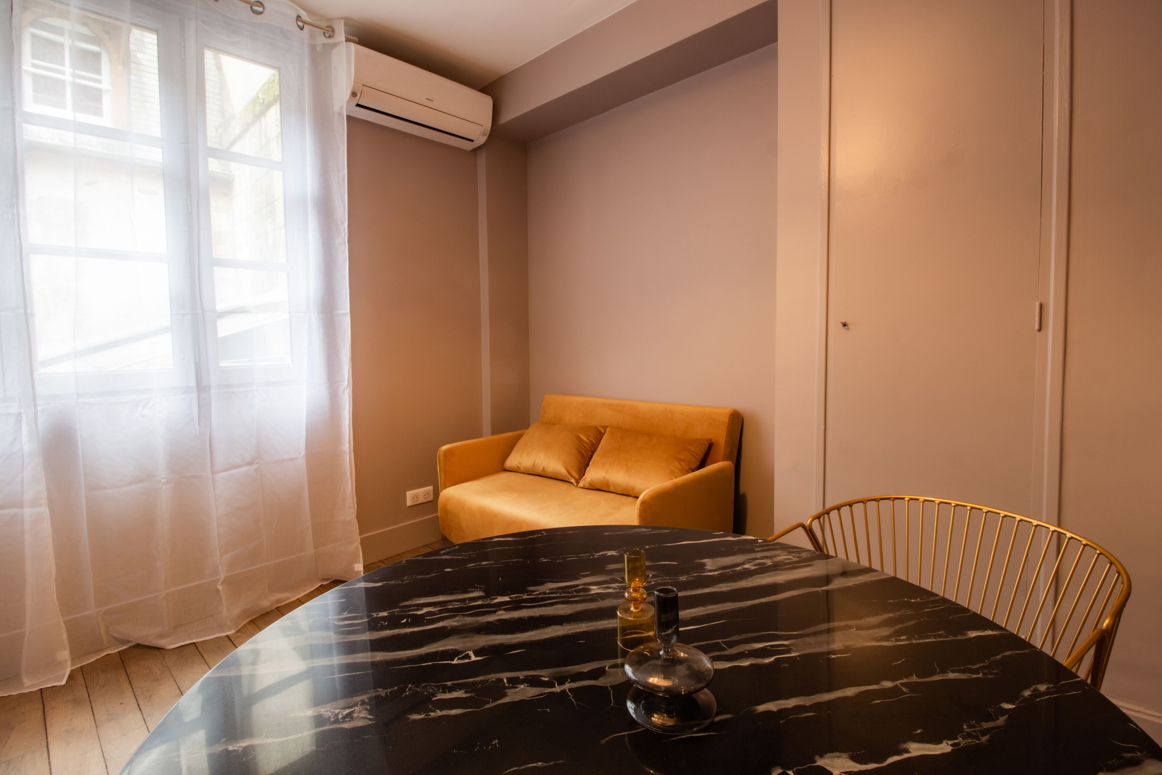 Una habitación con una mesa de mármol negro, una silla dorada, un sofá beige y una ventana con cortinas translúcidas.