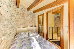 A bedroom with a stone wall, wooden beams, a bed with a polka dot bedspread, a small bedside table, and a bathroom visible through an open door.