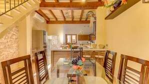 Un comedor con una mesa de vidrio, sillas de madera y un frutero en el centro. Se ve una cocina al fondo, con armarios de madera y una pared de piedra.