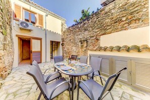 A stone-walled outdoor patio with a round table and chairs, a small table with a bottle and glasses, and a door leading to a building with wooden shutters and an air conditioning unit.