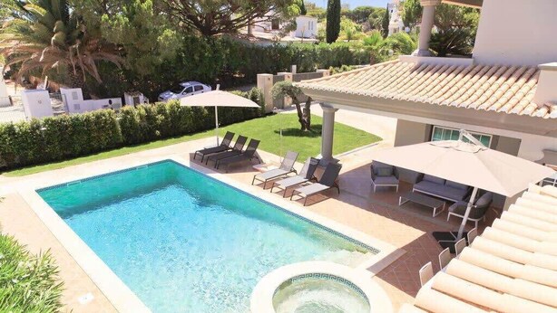 A swimming pool with lounge chairs and umbrellas, surrounded by a well-maintained garden and a building with a tiled roof. - Villa Selena - Beautiful Luxury villa with massage baths (Quinta do Lago)