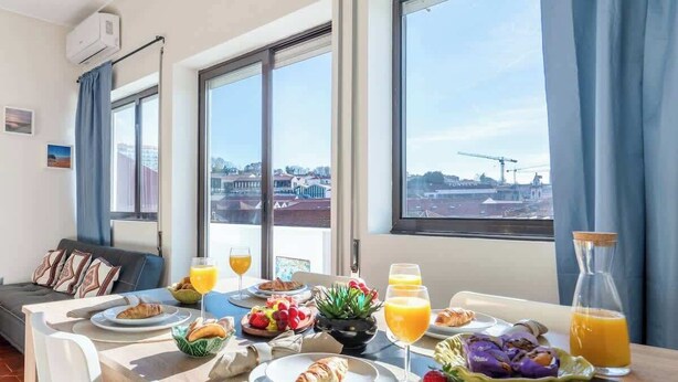 Una mesa de desayuno con cruasanes, frutas y jugo de naranja en una habitación luminosa con ventanas grandes.