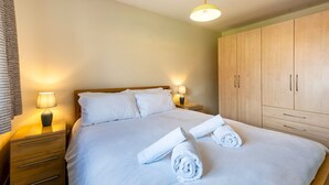 Un lit bien fait, avec du linge de lit blanc et deux serviettes enroulées, une table de chevet en bois avec une lampe, et une armoire en bois.