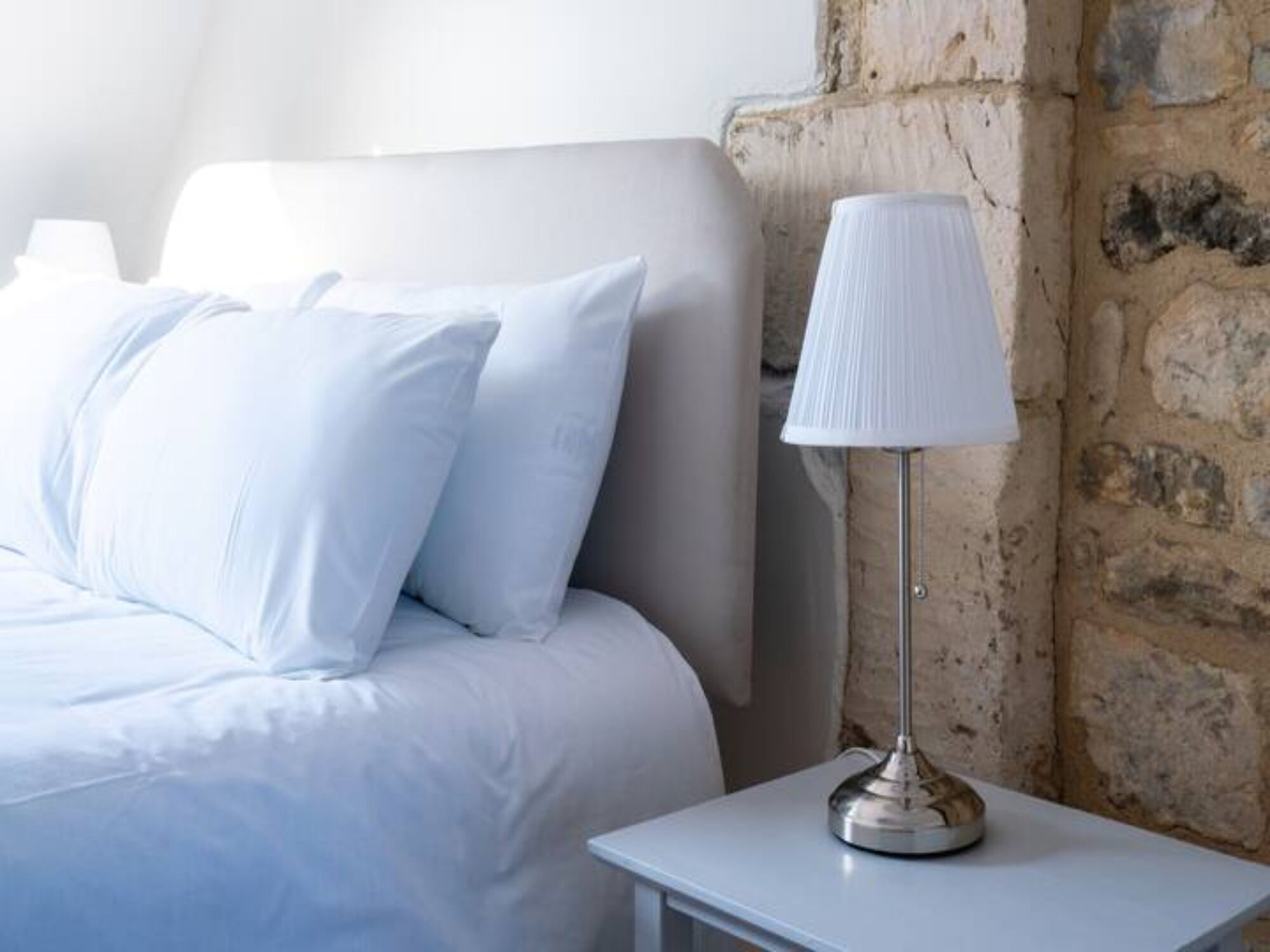 Ein Bett mit weißer Bettwäsche, ein Nachttisch mit Lampe und eine Steinwand.