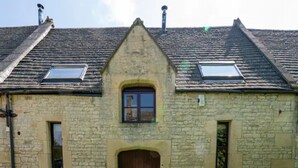 A stone building with a central entrance, two small windows on either side, and a dormer window above the entrance. - Converted 17th century barn with garden parking - Pass the Keys (Cheltenham)