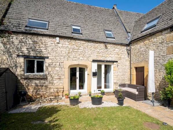 A stone building with a tiled roof, a covered patio with outdoor seating, and a small garden area with potted plants.