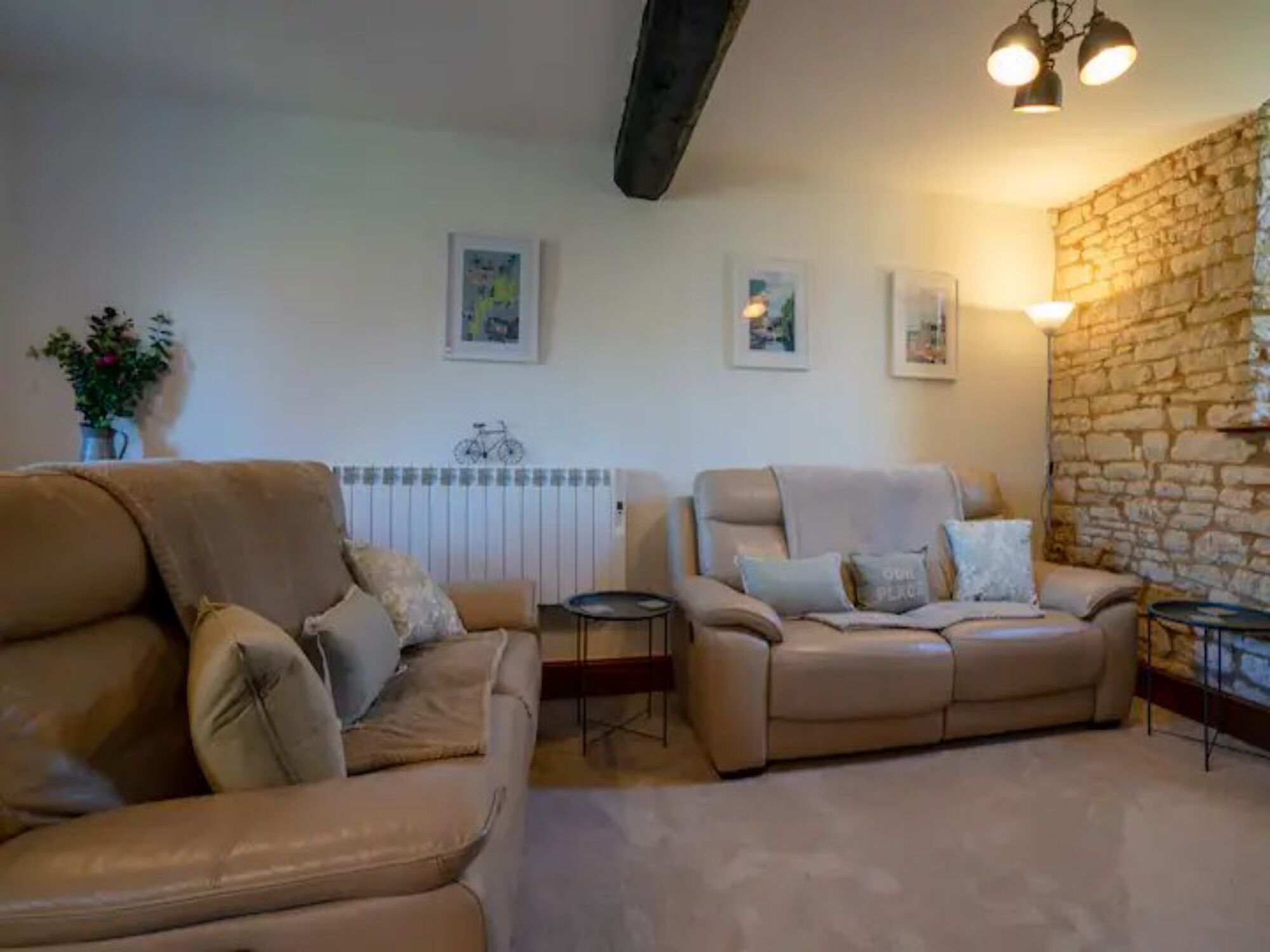 A living room with two beige sofas, a small round table, and a radiator. There are framed pictures on the wall and a potted plant on the left.