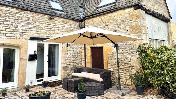 A stone building with a patio area featuring a white umbrella, wicker furniture, and potted plants. - Converted 17th century barn with garden parking - Pass the Keys (Cheltenham)