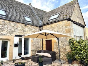 A stone building with a patio area featuring a white umbrella, wicker furniture, and potted plants.