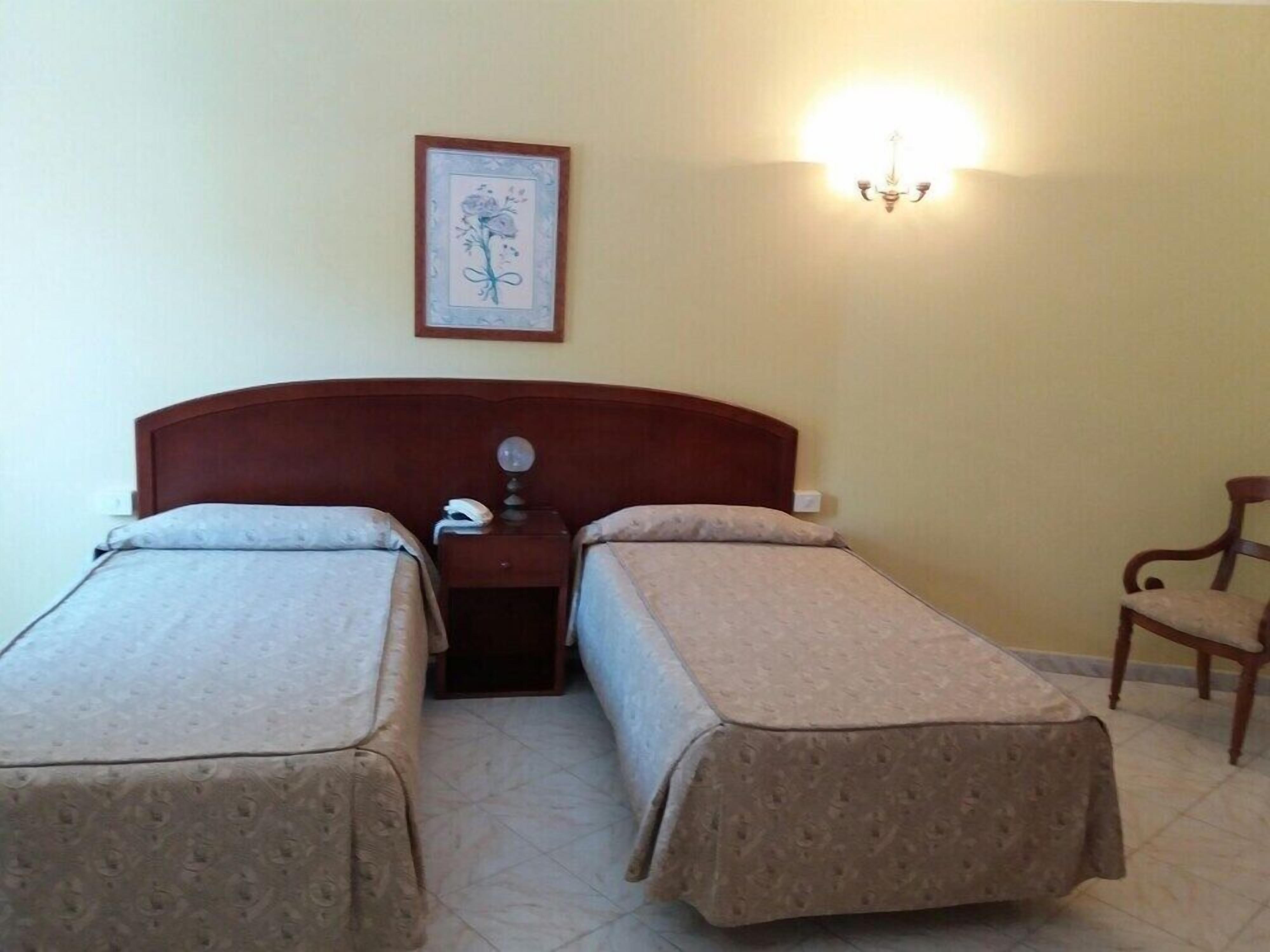 A room with two single beds, a nightstand with a lamp, a framed picture on the wall, and a chair.