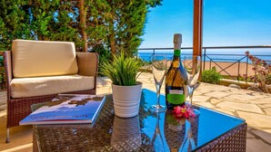 Una sedia in vimini, una pianta in vaso, una bottiglia di vino e un bicchiere su un tavolino di vetro con vista sul mare.