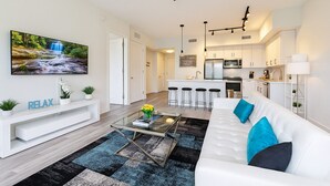 A modern living room with a white sofa, a coffee table, and a TV mounted on the wall. There is a kitchen area with white cabinets and a bar stool.