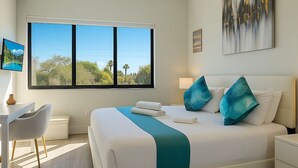 A modern hotel room with a large window, a bed with teal pillows, a desk, and a nightstand.