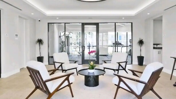 A modern interior with a round coffee table surrounded by white cushioned chairs, a ceiling light, and a view through glass doors to an outdoor area.