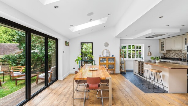 Une cuisine spacieuse avec une table à manger en bois, un îlot central intégré doté de tabourets et de grandes fenêtres offrant une vue sur le jardin.