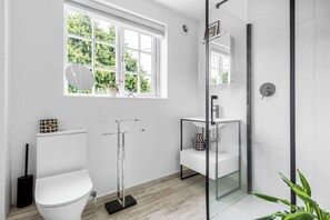 A modern bathroom with a white toilet, a glass shower enclosure, a white sink, and a window with a view of greenery. - Stunning 3-bed House in Saffron Walden (Saffron Walden)