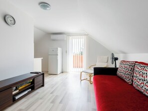 A modern, well-lit room with a wooden floor, a red sofa, a white chair, a small table with a remote control, and a clock on the wall. - Beautiful private villa for 9 guests with A/C, WIFI, TV, terrace and pets allowed (Keszthely)